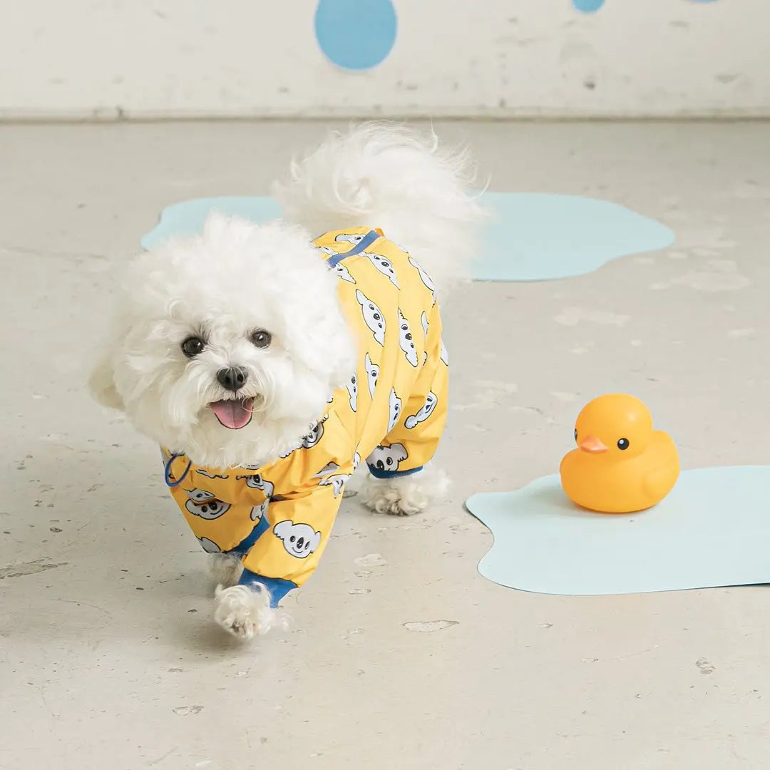 White dog wearing yellow pajamas with cartoon characters, standing next to a rubber duck on a light wooden floor.